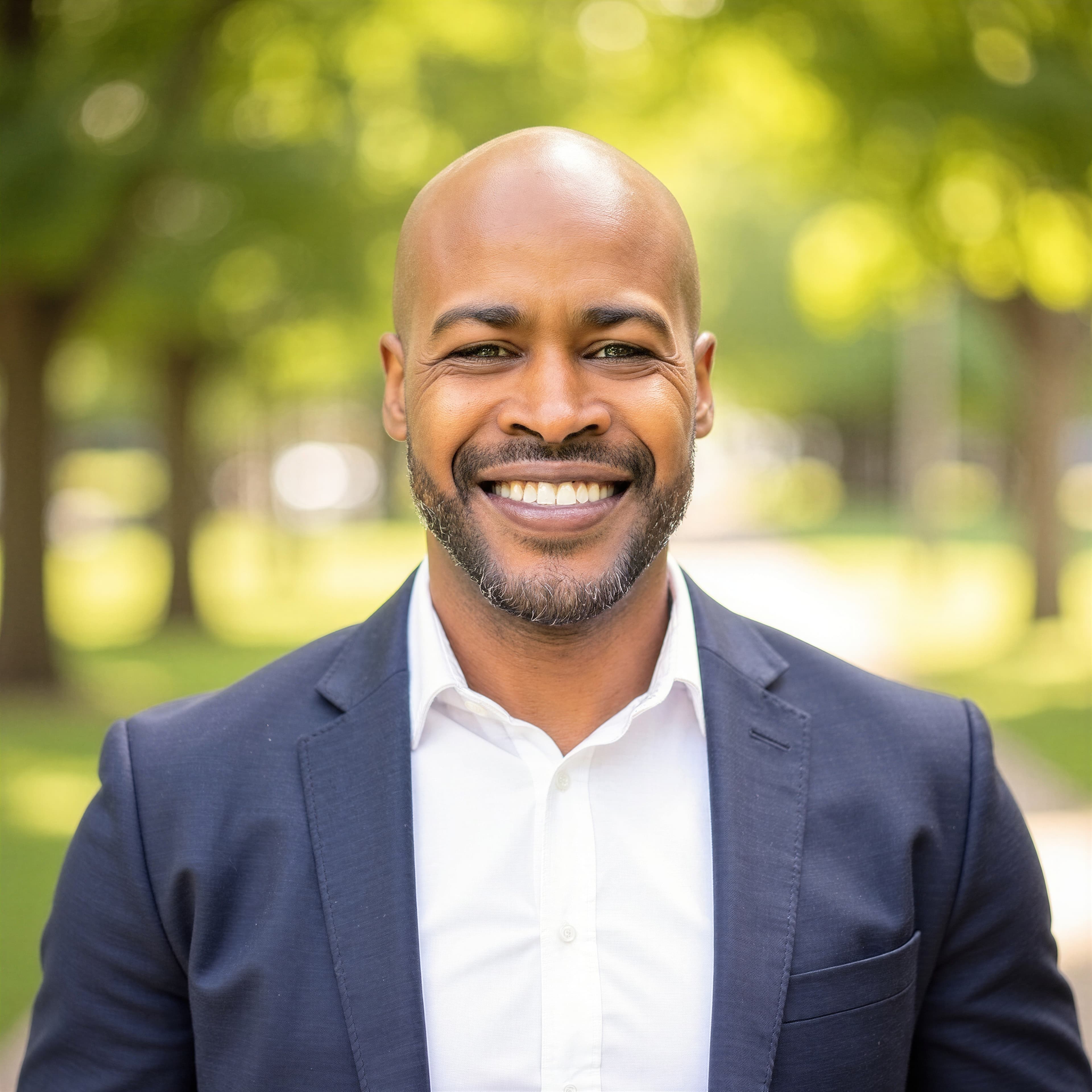 Smiling bald Black man with a beard wearing a navy blazer outdoors in a park.